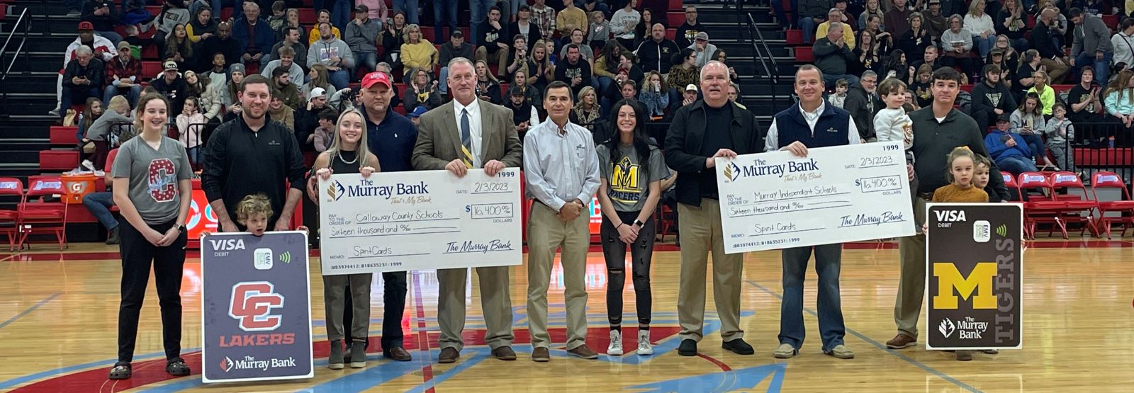 People holding large checks and debit card images in school gym.